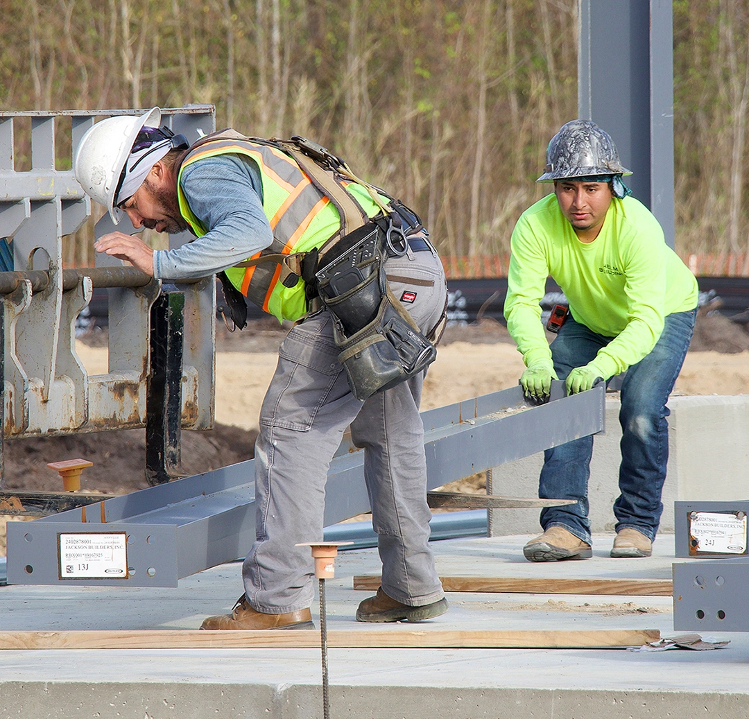 Two construction workers are assembling steel beams on a building site, with tools and safety gear visible. Trees are in the background.