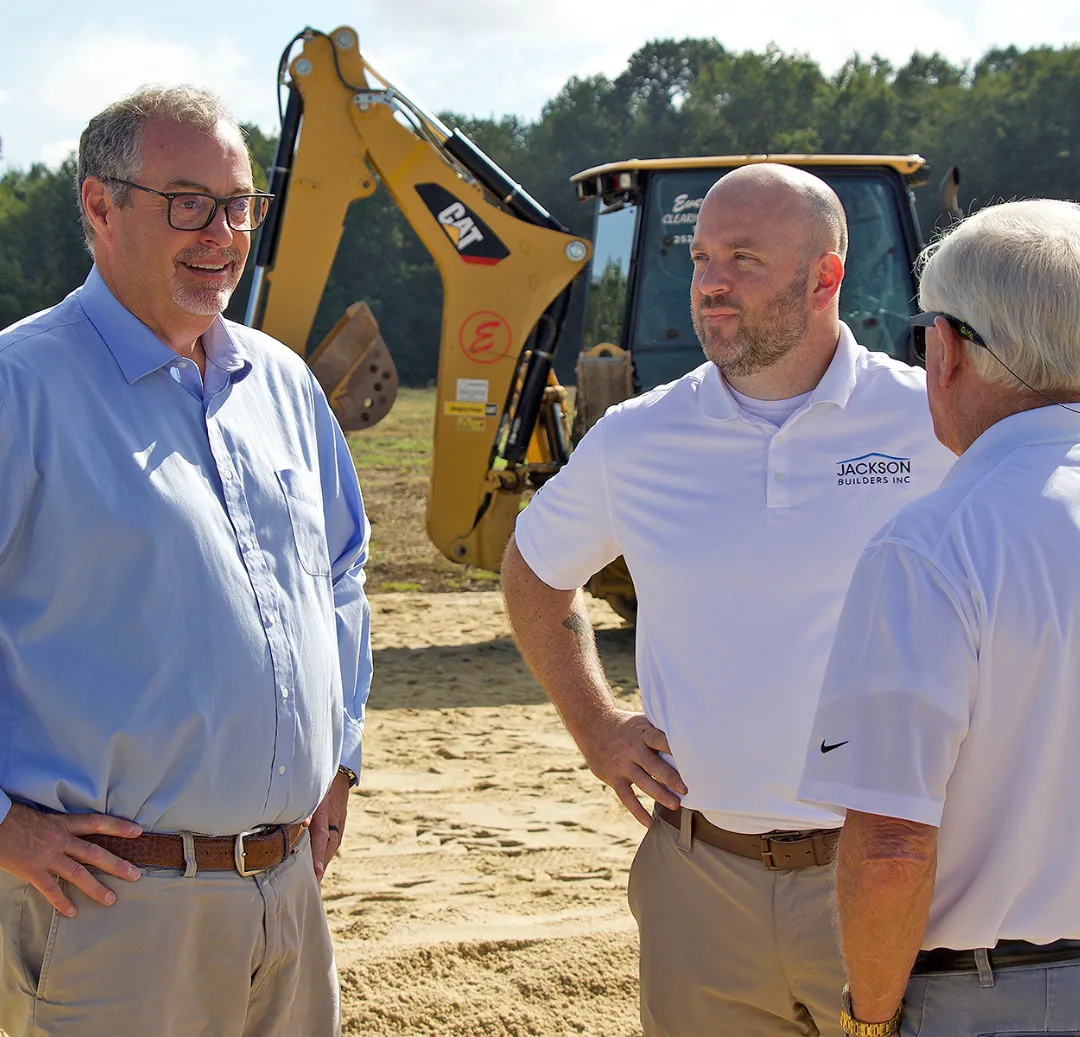 Three men in business attire engage in conversation at a construction site, with machinery and trees in the background